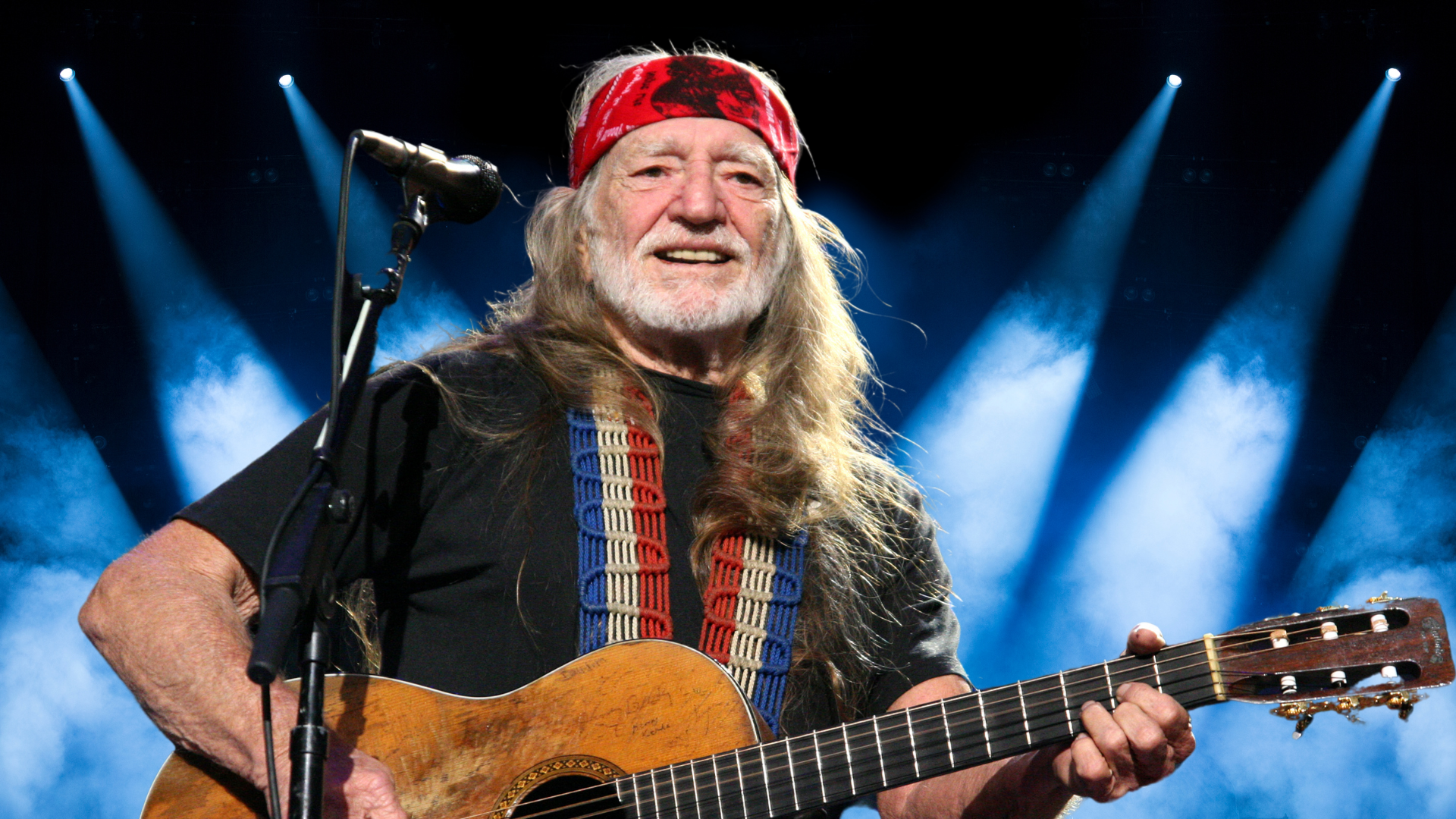 Willie Nelson holding a guitar and smiling on stage
