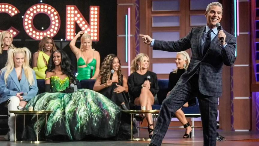 Andy Cohen speaks onstage during BravoCon alongside several Bravo reality television stars seated behind him.