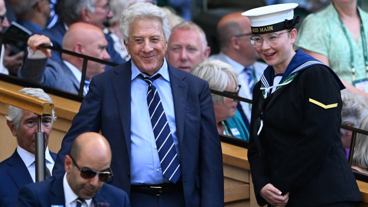 Dustin Hoffman stands in a blue suit and tie in the stands at Wimbledon.