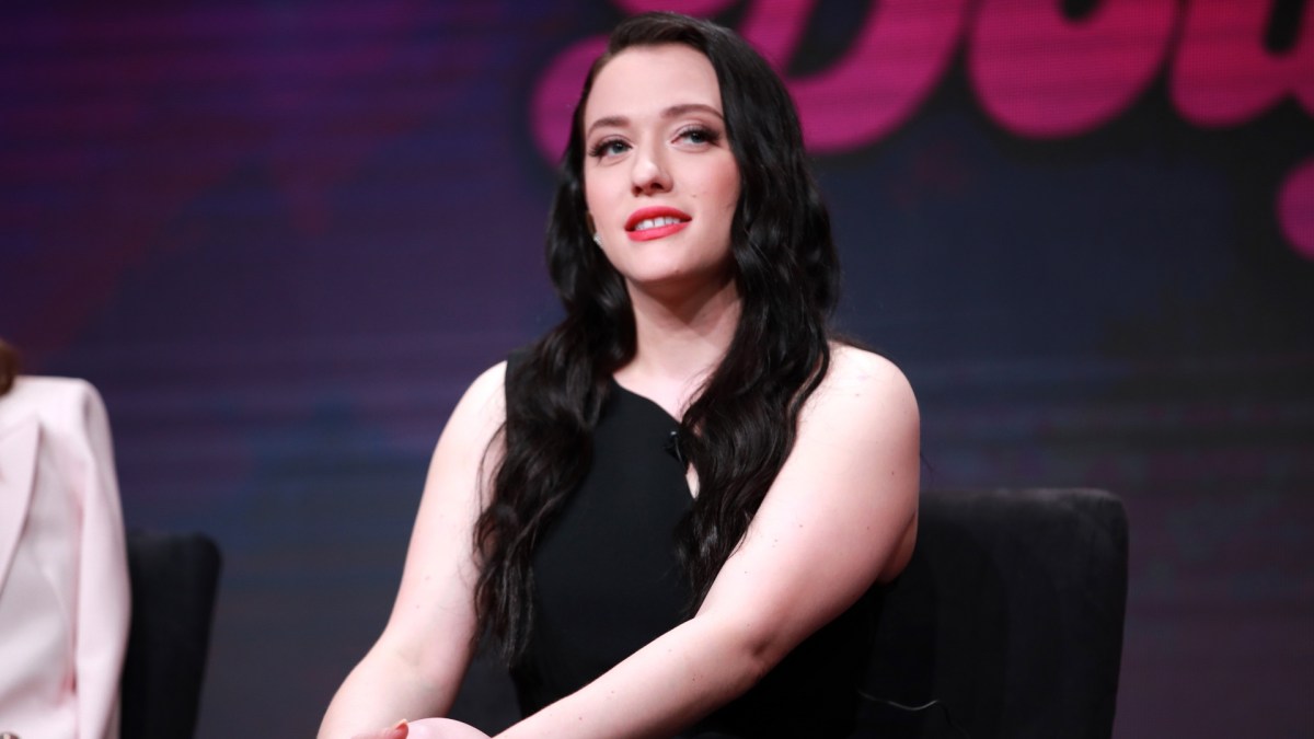 Kat Dennings wears a black dress and sits on stage.